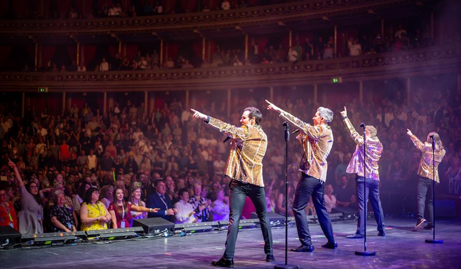 A photograph of musical theatre group Collabro performing on stage. The back of four men in gold jackets in a packed theatre. They each point to the s