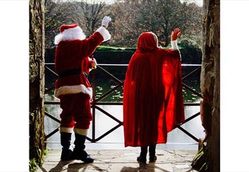 backs of father christmas and woman in red cloak waving out of castle archway.