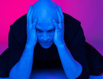 Photograph of a bald white man with his hands clasping the side of his head. the background and lighting is a hue of blue and pink.