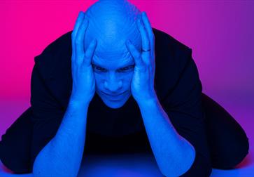 Photograph of a bald white man with his hands clasping the side of his head. the background and lighting is a hue of blue and pink.