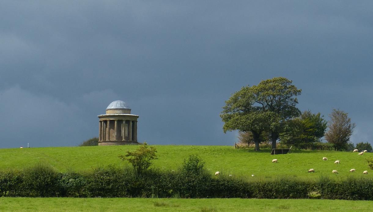 a photograph of a bright green grassy field from a distance, with dark grey moody sky. At the top of the feild is a round domed monument.