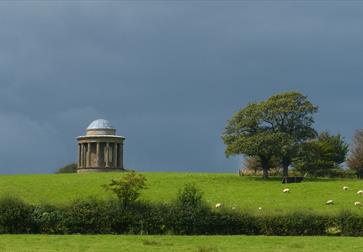 a photograph of a bright green grassy field from a distance, with dark grey moody sky. At the top of the feild is a round domed monument.