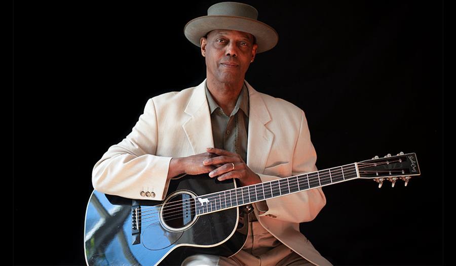 Photograph of balck man wearing a white suite jacket and brimmed hat with arms placed on his guitar. Against a black background.