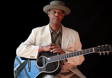 Photograph of balck man wearing a white suite jacket and brimmed hat with arms placed on his guitar. Against a black background.