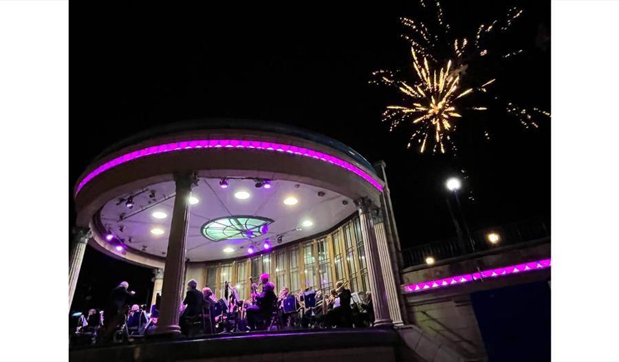 Image shows a band stand with a band playing on it surrounded by purple lights. There is a firework going off in the sky in the background. Image shows a band stand with a band playing on it surrounded by purple lights. There is a firework going off in the sky in the background.