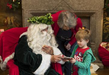 Man with long white hair, long beard and leaf headpiece giving a gift to a small boy.