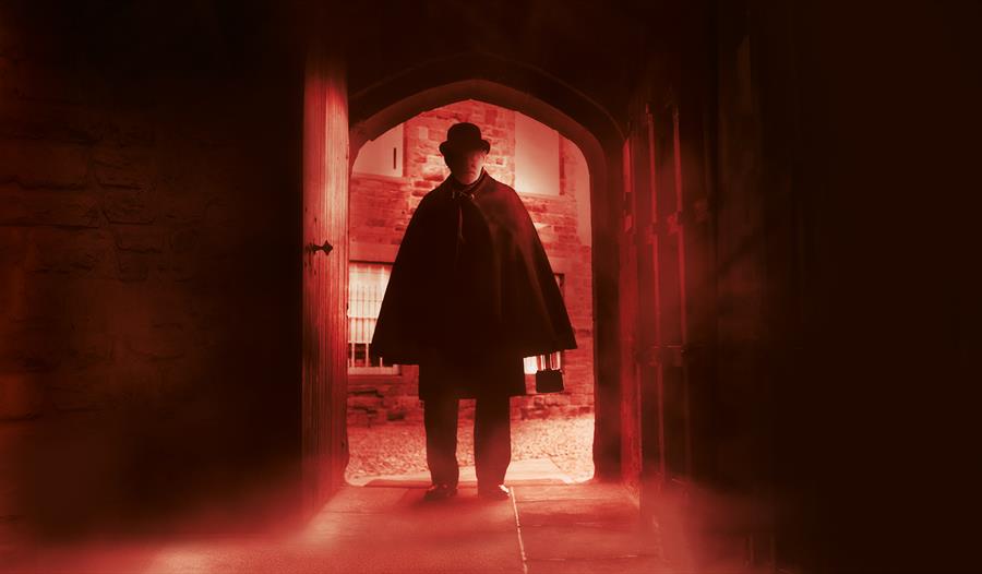 a dark photograph with a near silhouette of a man in a bowler hat and cloak as he enters through an arched wooden door. There is a red hue and a blur.