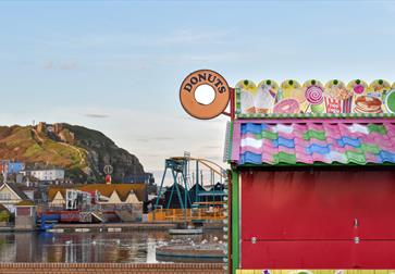 photograph of Hastings, with donut sign and colourful hut in the foreground and hill with funicular railway in the background.