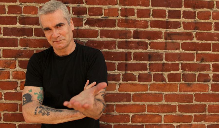 Photograph of white man in black t-shirt with tattoo arms standing against a brick wall.