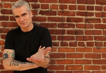 Photograph of white man in black t-shirt with tattoo arms standing against a brick wall.