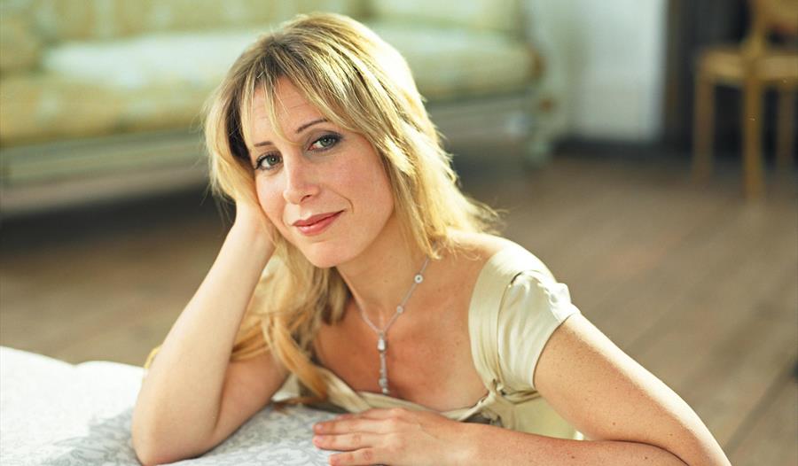 a photograph of a musician, taken from back of a sofa. A white woman with blond hair sits on floor and rests her elbows on seat of sofa, is smiling.