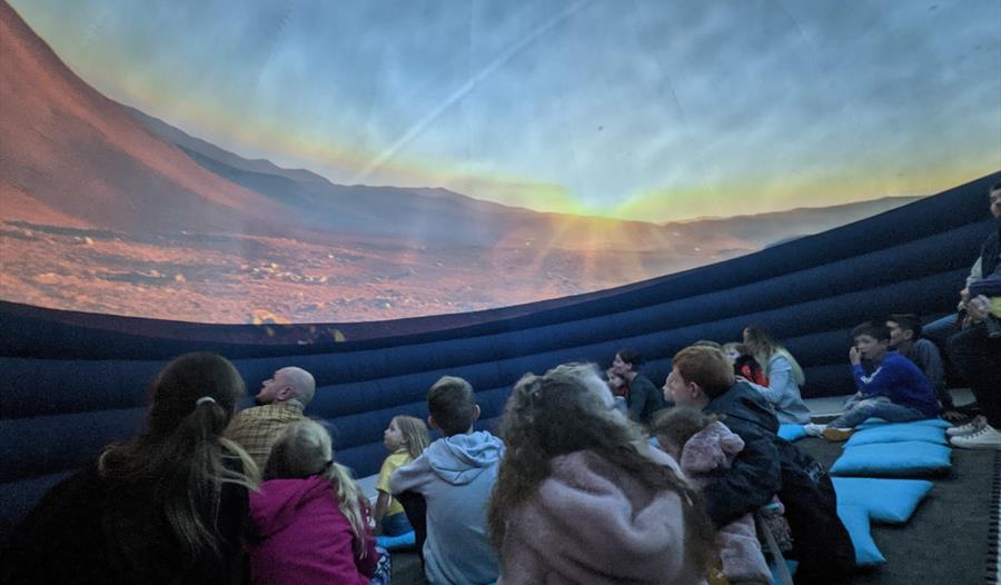 children stat inside a planetarium looking up a bare red planet landscape.