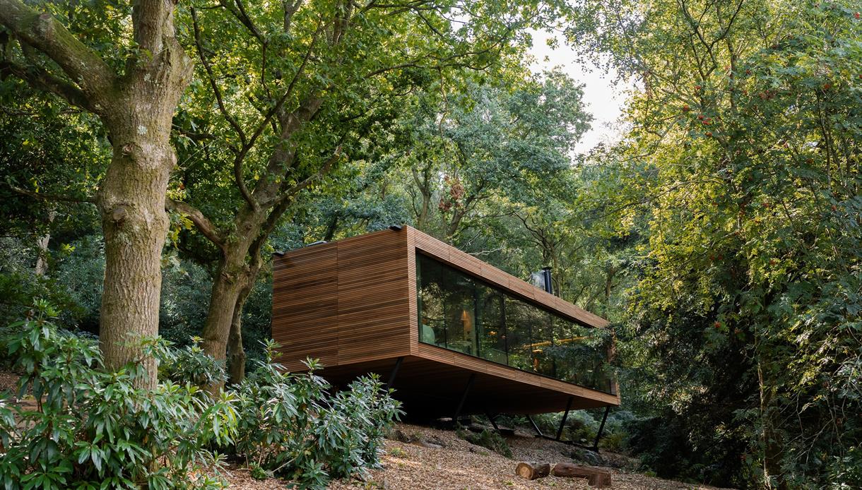 A suspended rectangular wooden lodge with large glass facade in a wooded landscape.