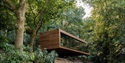A suspended rectangular wooden lodge with large glass facade in a wooded landscape.