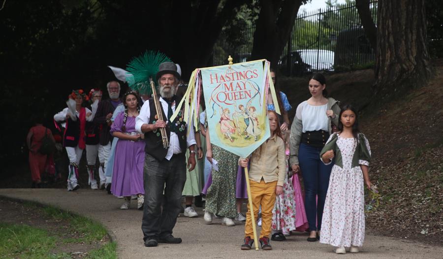 A May Queen procession with a child holding a banner and a chimney sweep at the front.