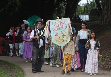 A May Queen procession with a child holding a banner and a chimney sweep at the front.