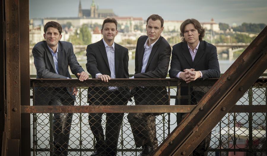 a photograph of four men standing against a rusty iron bridge. wearing suits and light coloured shirts.