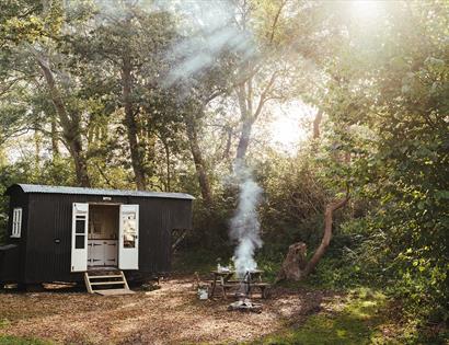 Photograph of a black glamping hut in a woods with smoking fire.