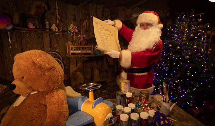 A photograph of Santa in his workshop holding up his list, a teddy bear in the foreground.