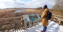A person wearing a brown coat and black woolly hat reading interpretation board, looking out towards a nature reserve with river. Decking underfoot is