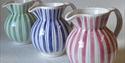 Three striped round ceramic jugs with handles in a row. The one farthest away is green and white, the middle is blue and white, the front one is pink