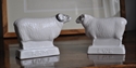 two small white ceramic sheep facing eachother on wooden furniture top. The bas of the left one, with horns, says 'ram'. The base of the right one say