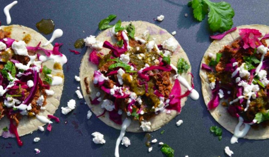 A photograph of three open tacos in a rowwith messy sour cream and coriander leaves over the grey surface. For Taco night at the hub bodiam rother