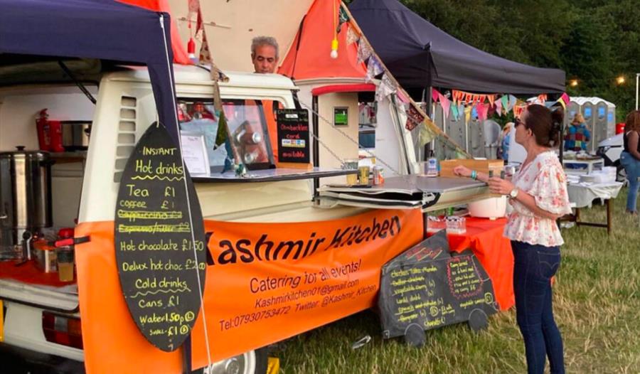a photograph of a food van in a field with lady standing to make her order. Van has orange banner saying Kashmir Kitchen.