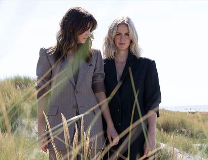 photograph of two women in grey and black suit jacket, one blonde and one brunette, standing on sand dunes amongst the grasses.