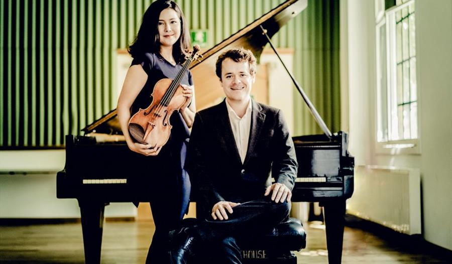 photograph of two musicians, a woman standing with violin and man seated with back against piano, green wallpaper in the background and light coming t
