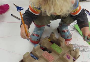 small blond boy in wellies standing on doodled paper with pencil in hand.