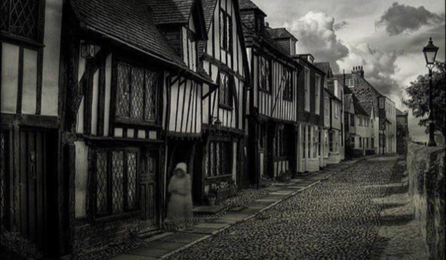 black and white photograph of a cobbled street with tudor houses and shadowy hooded figure