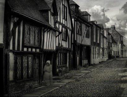 black and white photograph of cobbled street with single blurred ghostly figure walking beside tudor houses