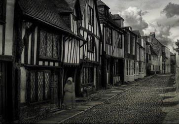 black and white photograph of cobbled street with single blurred ghostly figure walking beside tudor houses
