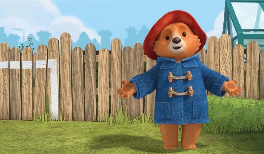 poster of cartoon bear in front of a garden fence wearing a blue duffle coat and a red hat.