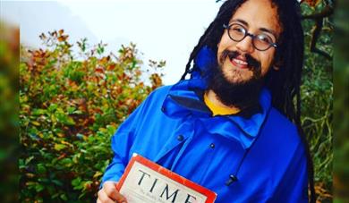 Rastafari historian Ras Benji stands in front of a hedge holding a copy of Time magazine. He has long dreadlocks and is smiling.