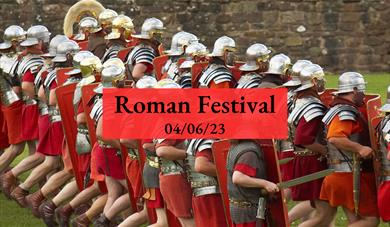 A group of men dressed in Roman centurion outfits marching along a field with the text 'Roman Festival 4/6/23' overlaid.