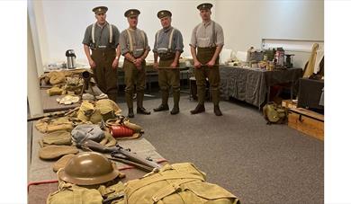 Men in First World War British Army uniforms stand behind tables with Great War military equipment, including webbing, helmets and packs