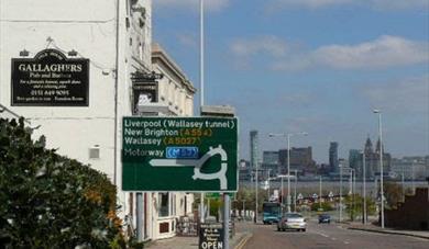 A street showing Woodside including cars and signs with the Liverpool skyline in the background.