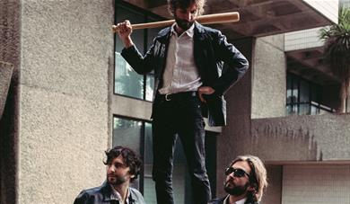 A man with a baseball bat stands on a wall above two other men leaning against the wall, looking relaxed and unaware