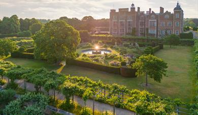 Aerial view of a historic mansion surrounded by manicured gardens. A fountain glows in sunlight, with lush trees and hedges under a serene sky.