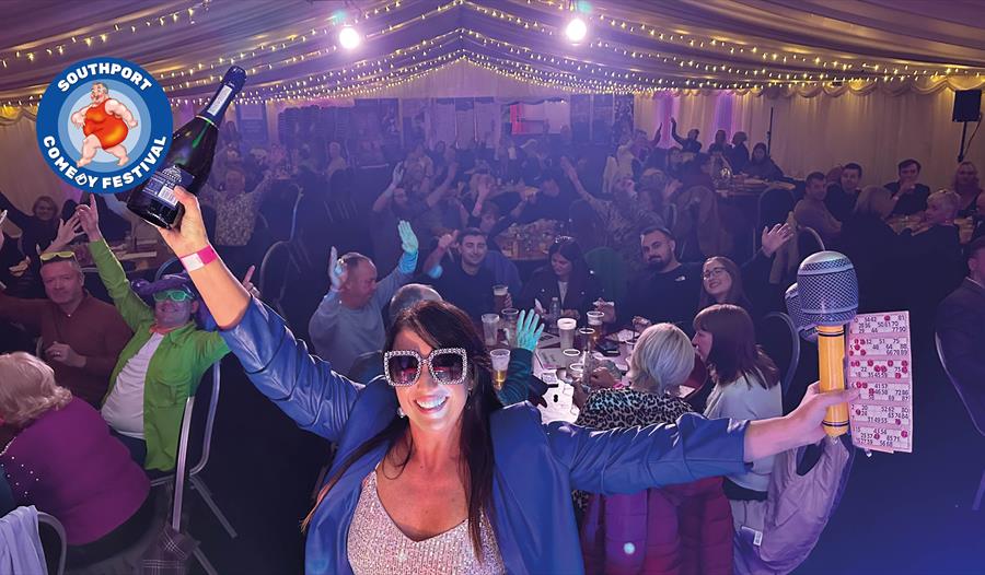 Image of a lady smiling wearing sunglasses and a sparkly top at Comedy Bingo holding a bottle of bubbly in one hand and a bingo card and blow up micro