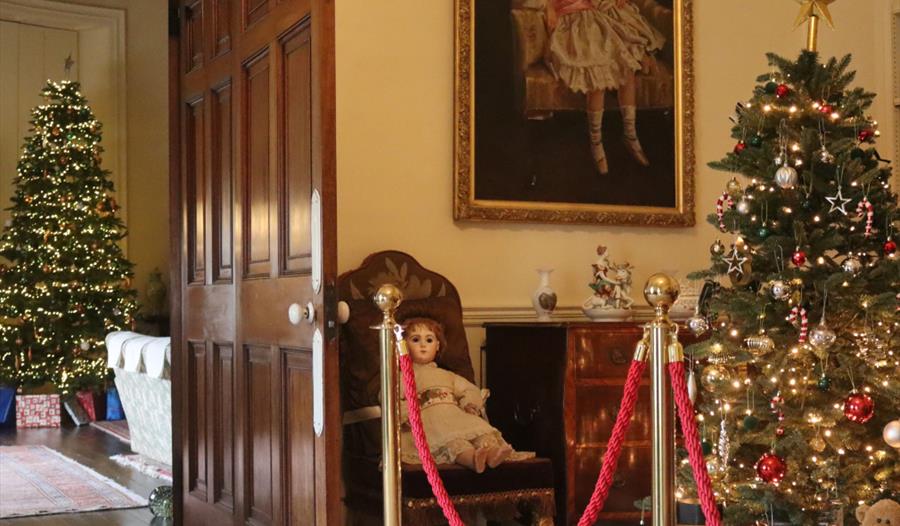 Christmas tree decorated with lights, a doll and a second Christmas tree in a room through a doorway.