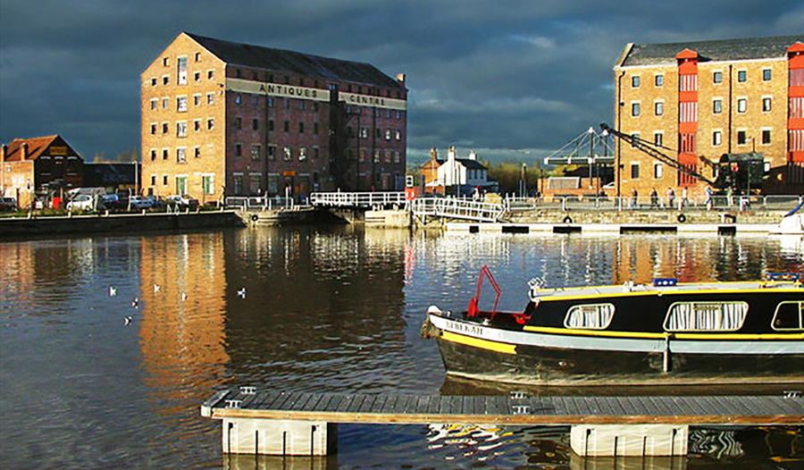 A colour photograph of Gloucester Docks including warehouse buildings, the water basin and a narrowboat in the foreground.