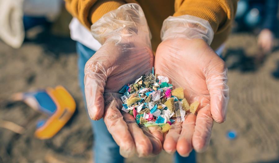Hands in clear gloves holding a pile of colorful microplastics on a sandy beach. The image conveys environmental concerns and pollution cleanup effort