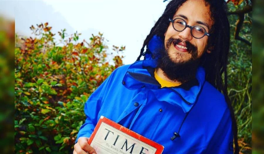 Rastafari historian Ras Benji stands in front of a hedge holding a copy of Time magazine. He has long dreadlocks and is smiling.