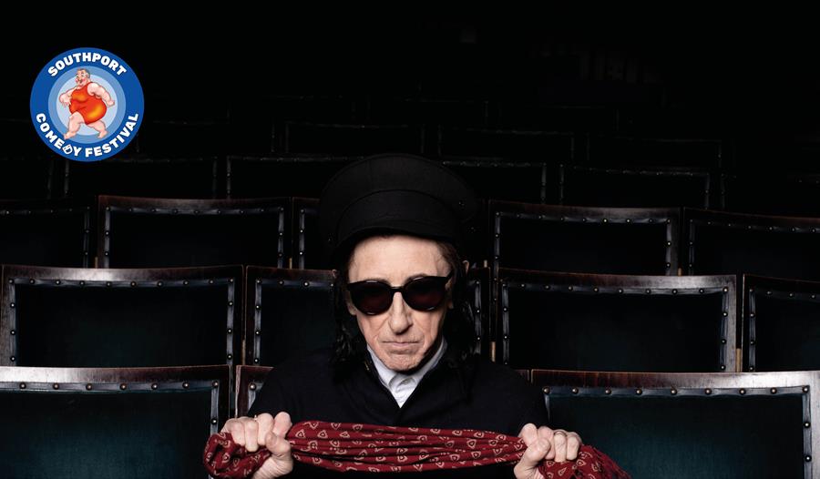 image of Dr John Cooper Clarke wearing dark sunglasses and a black hat sitting in an empty theatre looking to camera holding a red scarf