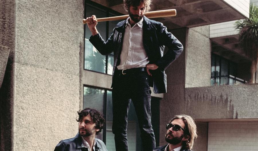 A man with a baseball bat stands on a wall above two other men leaning against the wall, looking relaxed and unaware