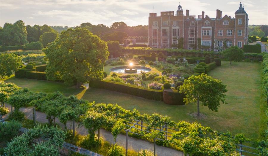 Aerial view of a historic mansion surrounded by manicured gardens. A fountain glows in sunlight, with lush trees and hedges under a serene sky.