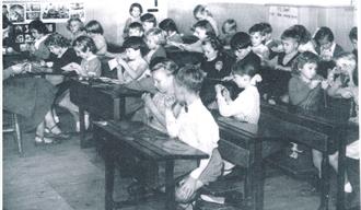 Black and white photograph of a school classroom. Children can be seen sitting at their desks working together.
